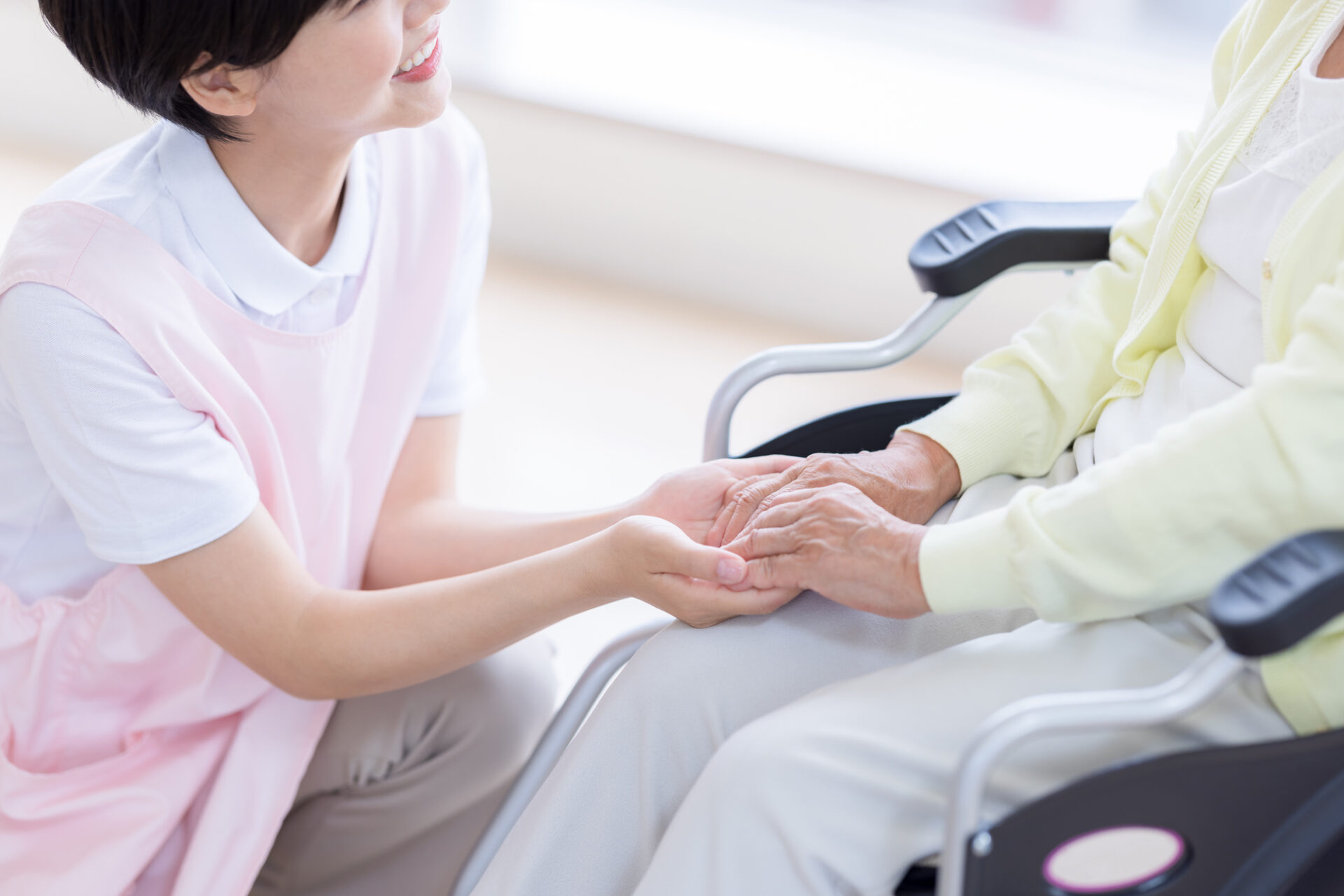 介護職員が高齢者の手を握りながらサポートする生活風景。車椅子での移動支援の様子。